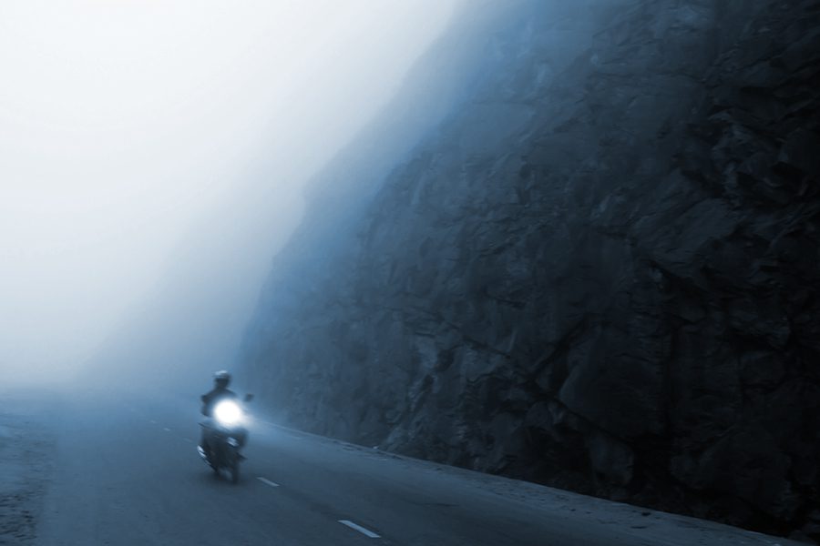 An unidentified male riding motorcycle on the dark misty mountain road at dusk.