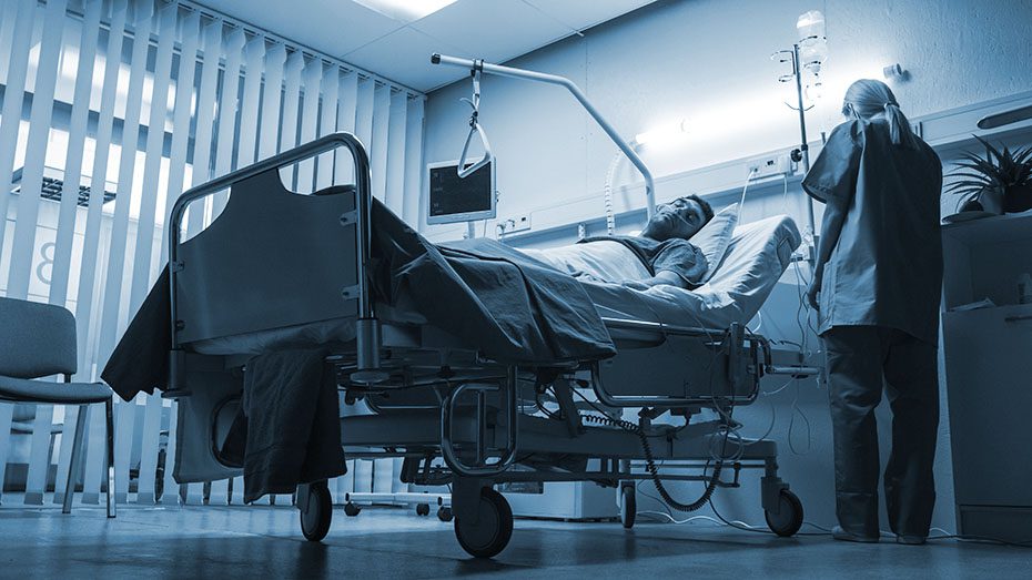 Man lays in hospital bed with nurse checking up on him. 