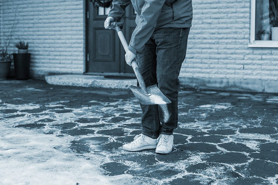 A man in blue jacket, jeans and white sneakers is using a shovel in front of his house