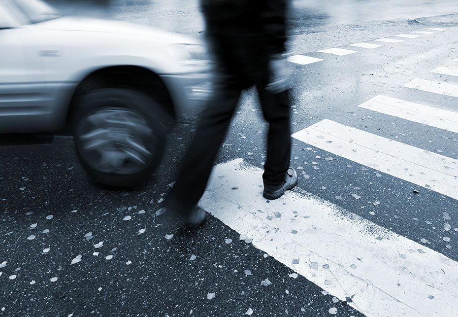 Man in black outfit walks across crosswalk while car makes a right turn. Motion blur